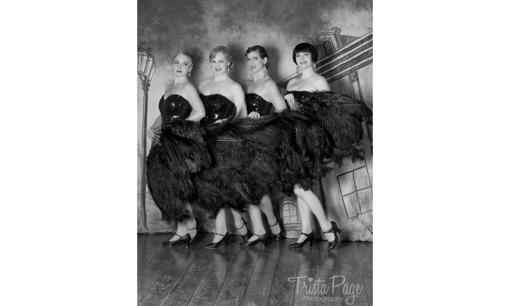 Group of dancers standing in a row with black feather fans and sequin tops..