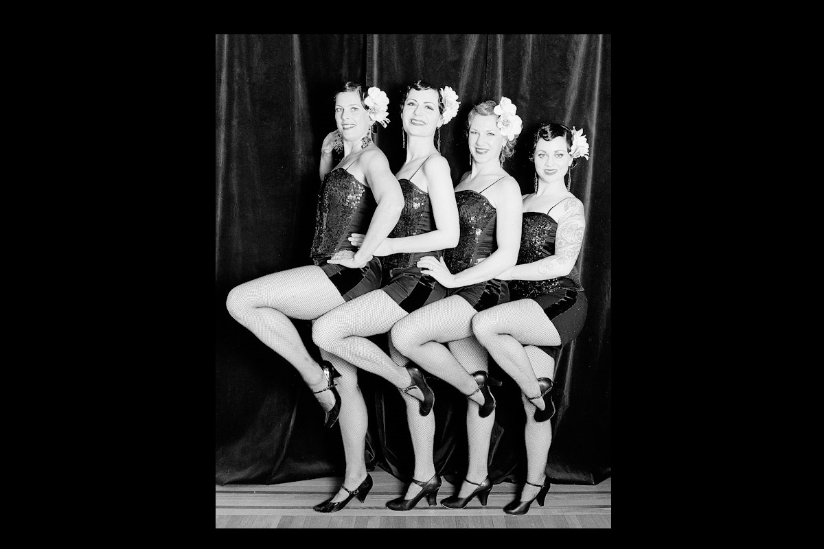 The Charleens dancers posing with one leg lifted with tuxedo shorts and sequin tops.